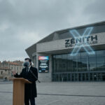 Un homme en costume derrière un pupitre devant le Zénith Sud de Montpellier sous un ciel gris et nuageux.