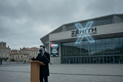 Un homme en costume derrière un pupitre devant le Zénith Sud de Montpellier sous un ciel gris et nuageux.