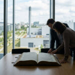 Un homme et une femme en tenue de bureau examinent un grand livre ouvert sur une table devant une fenêtre panoramique.