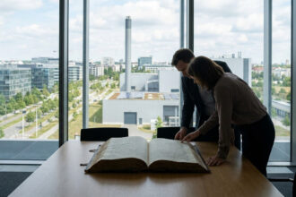 Un homme et une femme en tenue de bureau examinent un grand livre ouvert sur une table devant une fenêtre panoramique.