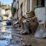 Une rue boueuse après une inondation avec des bottes en caoutchouc et une pelle couverte de boue contre un mur.