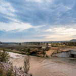 Un pont routier enjambant une rivière boueuse avec un ouvrage de décharge sous un ciel nuageux au crépuscule.