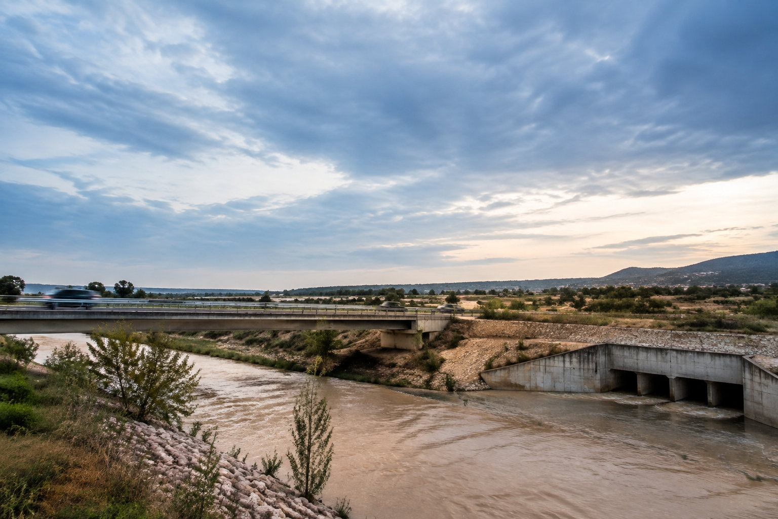 Un pont routier enjambant une rivière boueuse avec un ouvrage de décharge sous un ciel nuageux au crépuscule.